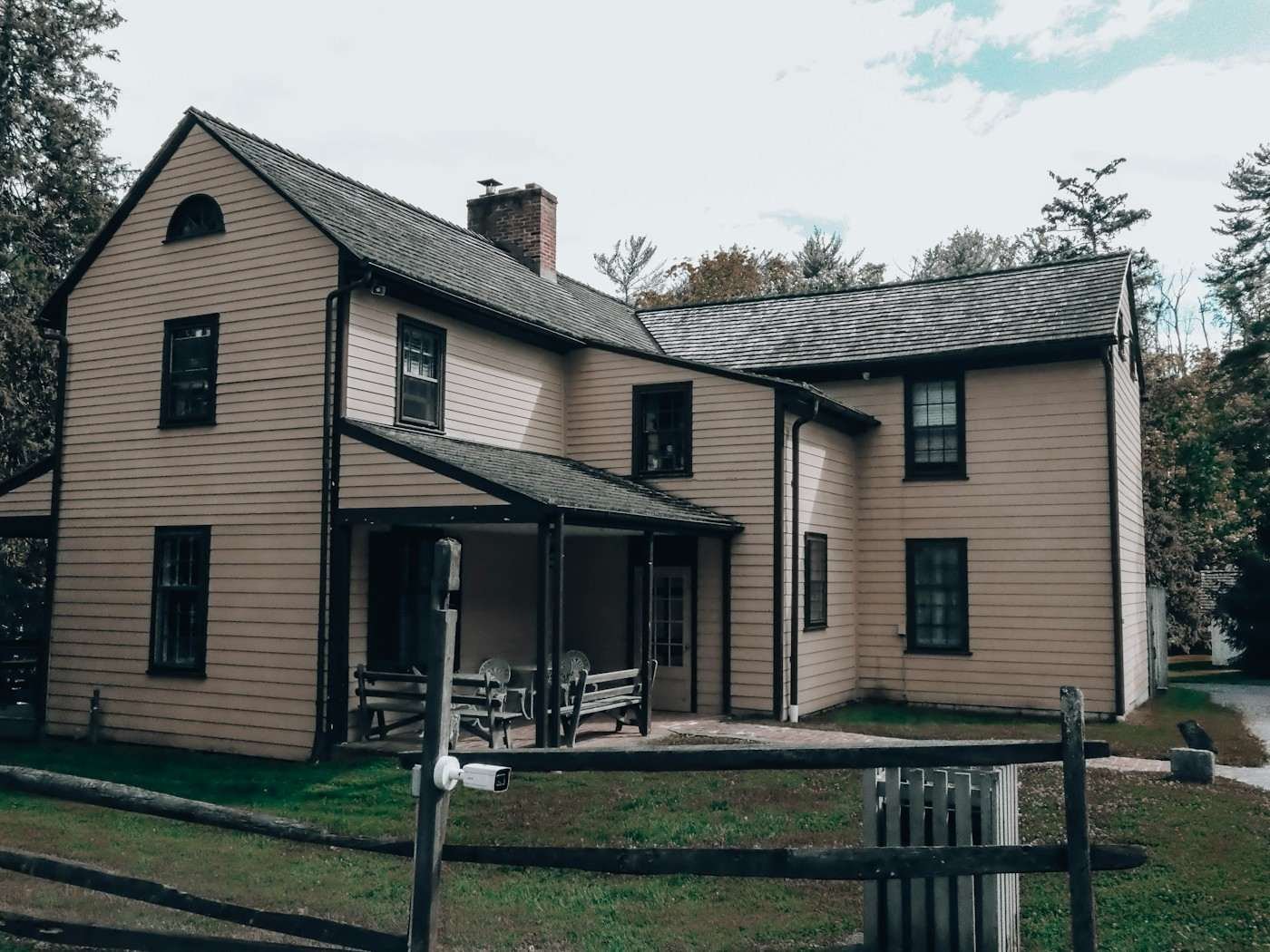 An older tan two-story house with a dark shingle roof and a wooden fence